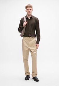 Camisa marrón de botones, pantalones beiges y zapatos negros. El modelo sostiene un bolso de hombro gris, con una textura suave y un diseño minimalista.