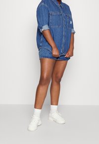 Jeansskjorta med uppvikta ärmar, två framfickor; i kombination med jeansshorts. Modellen har på sig vita sneakers och ankelsockor.