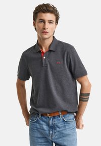 Graues Polo-Shirt mit rotem Innensaum am Kragen, kurzen Ärmeln, einer zwei-knöpfigen Knopfleiste und einem roten, gestickten Logo auf der Brust. Getragen mit blauen Jeans.