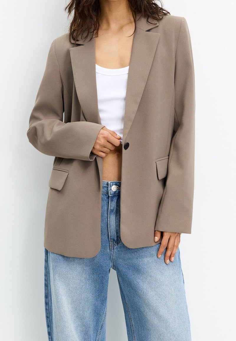 Femme portant un blazer beige oversize, un débardeur court blanc et un jean taille haute bleu clair, debout sur fond blanc.