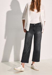 Black wide-leg jeans with light denim texture, paired with a white tank top and a loose white shirt. Worn with beige sneakers featuring green accents.