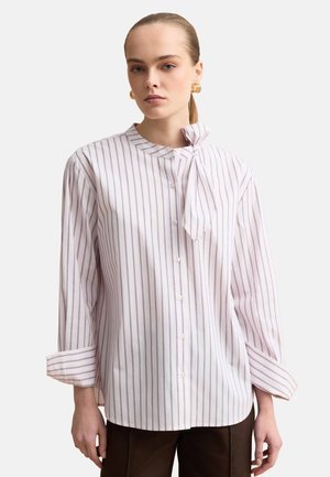 Mujer con blusa blanca de delgadas rayas verticales y un lazo en el cuello lateral, combinada con pantalones oscuros y pendientes de aro dorados.