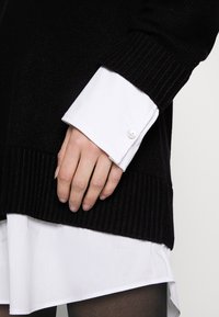 Maglione nero lavorato a maglia con polsini a coste, indossato sopra una camicia bianca con colletto. La mano poggia sul bordo dell'orlo del maglione.