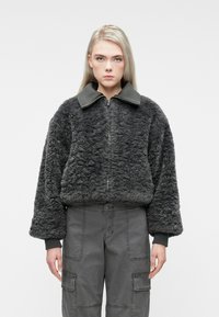 Ung person med langt blondt hår iført en mørkegrå struktureret zip-up jakke og grå cargo bukser, stående mod en hvid baggrund.