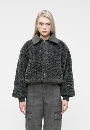 Ung person med langt blondt hår iført en mørkegrå struktureret zip-up jakke og grå cargo bukser, stående mod en hvid baggrund.