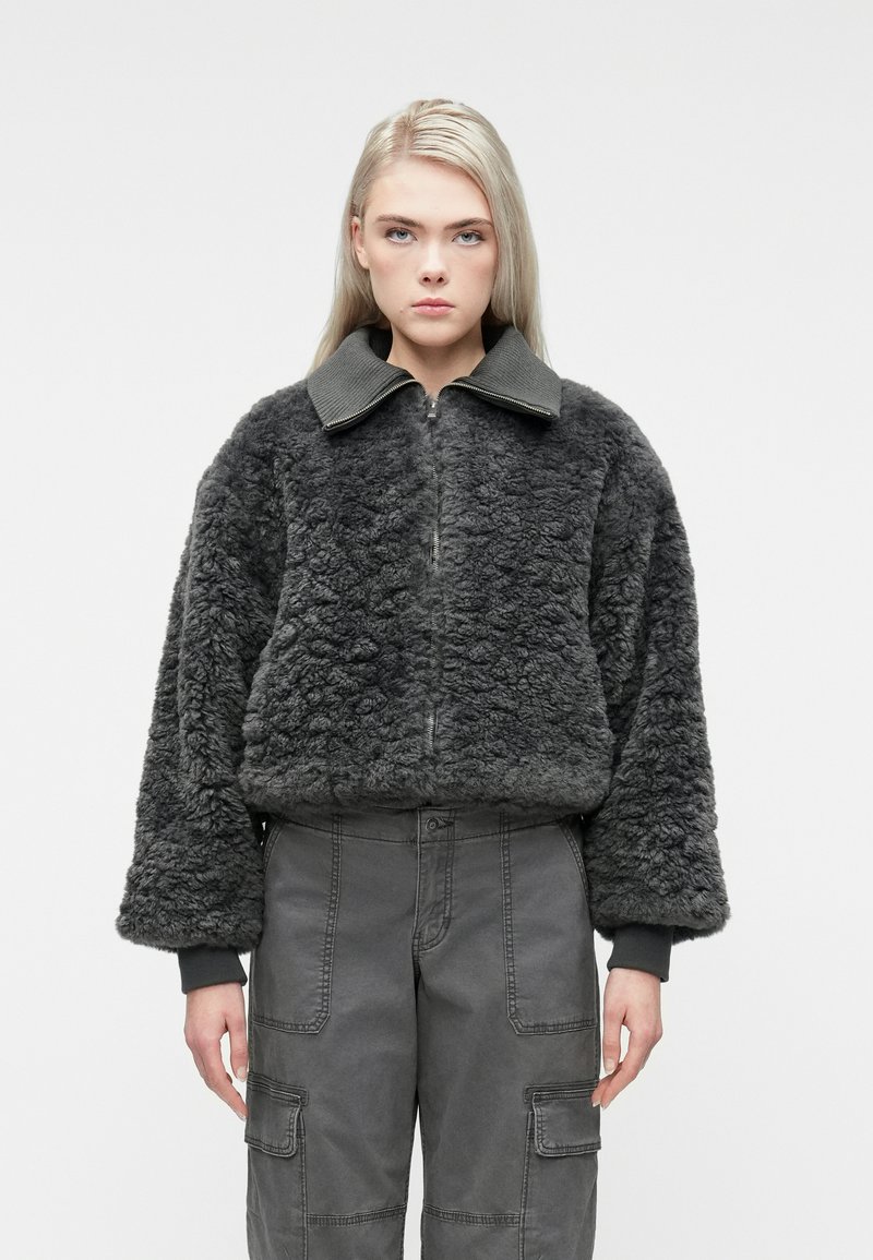 Ung person med langt blondt hår iført en mørkegrå struktureret zip-up jakke og grå cargo bukser, stående mod en hvid baggrund.