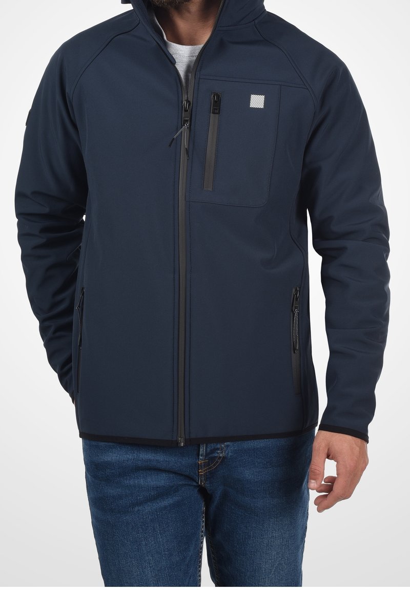 Homme portant une veste zippée bleu marine avec poches zippées sur la poitrine et les côtés, par-dessus une chemise blanche et un jean bleu.