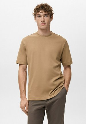 Jeune homme aux cheveux bouclés portant un t-shirt beige à manches courtes et un pantalon foncé, debout avec une main dans la poche devant un fond uni.
