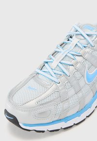 Sko med lätt grå mesh och syntetiska material, accentuerad med blå snören och detaljer. Noterbara texturerade överlägg och dämpad sula.