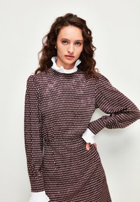 Robe à motifs marron avec col et poignets en dentelle blanche, dotée de manches longues et d'une taille ajustée. Le design comprend des formes verticales.