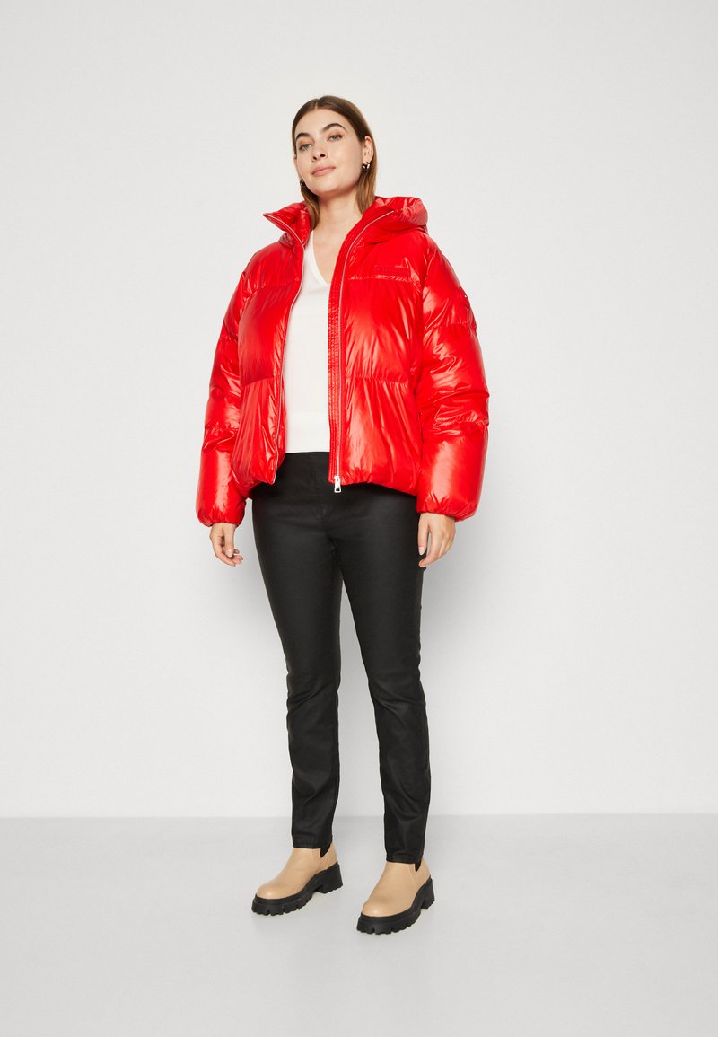 Donna in piedi con giacca a vento rossa, top bianco, pantaloni neri e stivali beige su uno sfondo bianco semplice.