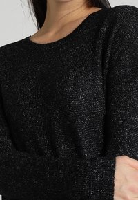 Zwarte trui met een gestructureerd breipatroon, losse pasvorm en een ronde halslijn. De stof bevat metallic draden voor een subtiele glans.