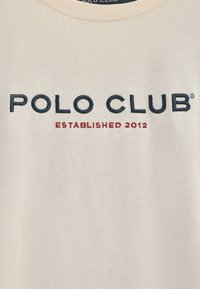 Beżowa bawełniana koszulka z haftem "POLO CLUB" w granatowym kolorze oraz czerwonym napisem "ZAŁOŻONA W 2012" poniżej. Standardowy okrągły dekolt.
