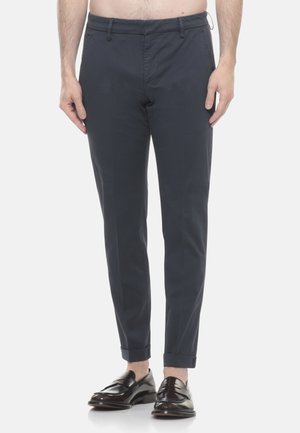 Uomo che indossa pantaloni aderenti blu navy con orli risvoltati e mocassini in pelle nera, in piedi davanti a uno sfondo bianco.