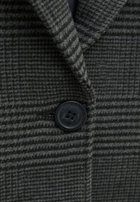 Manteau en laine gris foncé avec un motif à carreaux subtil et un bouton noir à quatre trous unique fermé près du col.