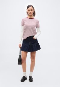 Νεαρή γυναίκα με σγουρά μαλλιά, που φοράει ριγέ κοντομάνικη μπλούζα πάνω από λευκά μακριά μανίκια, τζιν φούστα, λευκές κάλτσες, μαύρα μοκασίνια, κρατώντας μαύρη τσάντα χειρός.