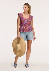 Góra peplum w intensywnym różowym kolorze z wielokolorowym kwiatowym wzorem, zestawiona z jasnymi szortami jeansowymi. Modelka trzyma szerokokątny kapelusz ze słomy, ma na sobie niebieskie sandały.