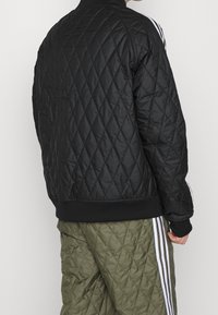 Svart vadderad bomberjacka med ribbade ärmslut, en rund krage och vita randiga detaljer längs ärmarna. Bärs med matchande gröna vadderade byxor.