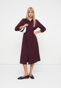 Burgunderfarget knappingkjole med belte i midjen, lange ermer og avslappet passform; kombinert med svart og hvitt håndbagasje og blanke, mørke flater.