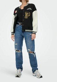 Czarna i biała kurtka varsity z naszywkami, noszona na czarnej bluzce, w połączeniu z zniszczonymi niebieskimi dżinsami i szarymi trampkami.