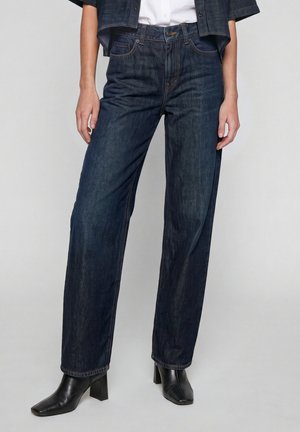 Donna che indossa jeans blu scuro a gamba dritta, stivali neri con tacco e una giacca di jeans corta sopra una camicia bianca, in piedi davanti a uno sfondo uniforme.