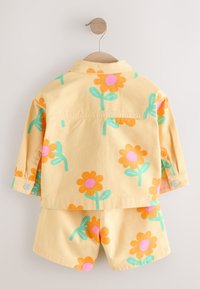 Ensemble jaune pour tout-petit avec des imprimés de fleurs orange et rose, composé d'une veste à manches longues et d'un short assorti sur un cintre en bois.