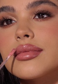 Błyszczyk w przezroczystej tubce, nałożony na pełne usta. Usta są błyszczące, w odcieniu nude różu, o gładkiej teksturze. Naturalny makijaż podkreśla rysy twarzy.