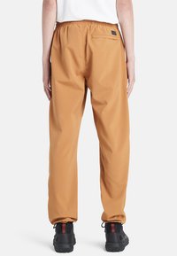 Waterbestendige tan kleurige broek met een elastische tailleband, achterzak en een tappered design; gecombineerd met gestructureerde zwarte schoenen met rode accenten.