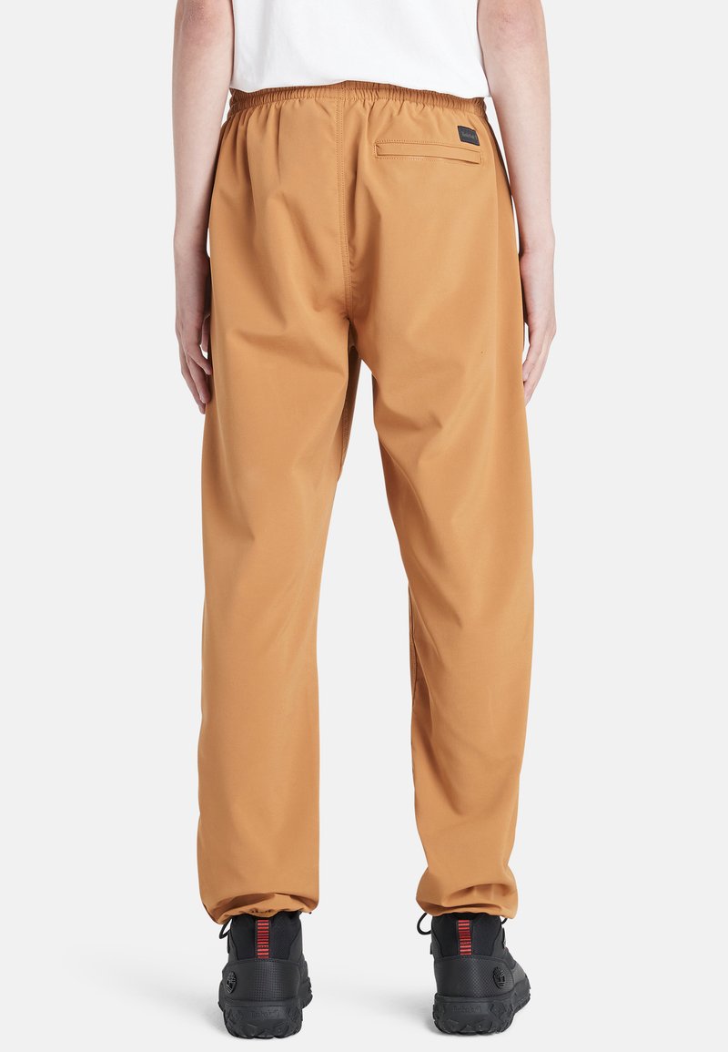 Wasserabweisende beige Hose mit elastischem Bund, Gesäßtasche und tailliertem Schnitt; kombiniert mit strukturierten schwarzen Schuhen mit roten Akzenten.