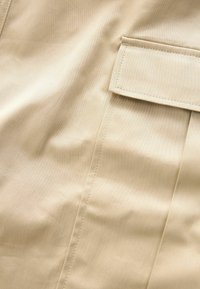 Détail de tissu beige montrant un rabat de poche rectangulaire fermé avec des surpiqûres visibles sur un matériau lisse.