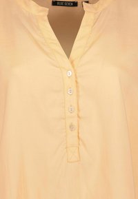Blusa gialla chiaro con scollatura a V, caratterizzata da cinque bottoni bianchi sul davanti e dettagli di cucitura sottili lungo il collo e il patta.