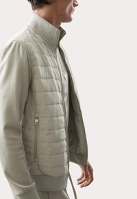 Giacca imbottita di un grigio chiaro con colletto alto e chiusura frontale con zip. Presenta polsini a coste e una finitura del tessuto liscia. Tasca laterale con zip visibile.