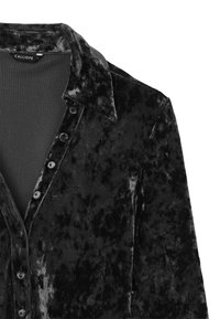 Camicia in velluto nero con colletto, chiusura frontale con bottoni e superficie leggermente strutturata. Presenta una fodera interna liscia. Etichetta del marchio visibile.