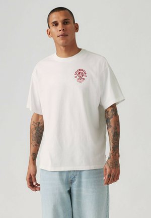 Tatuoitu mies käyttää valkoista Levi'sin T-paitaa, jossa on punainen logo, ja vaaleansinisiä farkkuja yksivärisen vaalean taustan edessä.