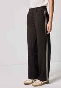 Persona che indossa pantaloni neri a gamba larga con strisce laterali beige e sneakers beige, in piedi contro uno sfondo chiaro e semplice.