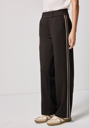 Persona che indossa pantaloni neri a gamba larga con strisce laterali beige e sneakers beige, in piedi contro uno sfondo chiaro e semplice.