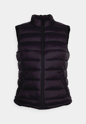 Person trägt eine schwarze gesteppt gefütterte Weste, die über einem schwarzen Strickpullover mit blauen Jeans hochgezogen ist.