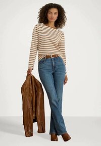 Nainen, jolla on päällä raidallinen pitkähihaisuus paita, siniset farkut ja ruskeat saappaat, pitää ruskeaa nahkatakkia valkoista taustaa vasten.