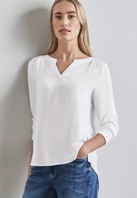 Weiße Bluse mit langen Ärmeln und V-Ausschnitt, die einen lockeren Schnitt, weiche Textur und seitliche Schlitze aufweist, kombiniert mit blauen Denim-Jeans.
