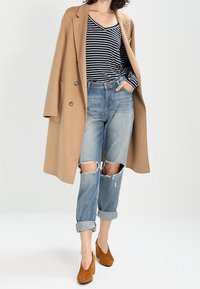 Cappotto beige a doppio petto, maglietta a righe bianco e nera, jeans blu strappati con risvolti, e scarpe con tacco alte marroni.