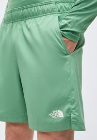 Πράσινο αθλητικό σορτς με ελαστική μέση, πλαϊνή τσέπη με χέρι μέσα και λευκό λογότυπο The North Face κοντά στο τελείωμα.