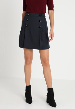 Mini-jupe bleu marine avec fermeture à boutons double boutonnage, poches avant plissées et texture lisse. Portée avec des bottines noires.