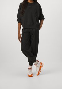 Felpa nera con scollo rotondo e logo ricamato, abbinata a pantaloni coordinati. Sneakers bianche e arancioni completano l'outfit.