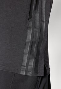Schwarzes Sportoberteil mit strukturiertem Stoff und glatten schwarzen Streifen an den Ärmeln. Seitenschlitze erhöhen die Flexibilität und den Komfort.