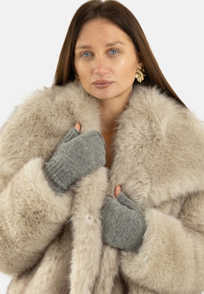 Jasnobeżowy płaszcz z futrzanym futerkiem z dużym kołnierzem, wyposażony w szare dziergane rękawiczki z otworem na kciuk, podkreślający teksturę i warstwowy design.