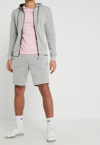 Sudadera gris con cremallera y cordones, camiseta rosa clara, pantalones cortos grises, calcetines blancos y zapatillas blancas. Tejido suave, corte atlético.
