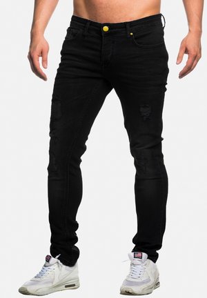 Mann trägt schwarze zerrissene Skinny-Jeans und weiße Nike Air Max Sneaker, steht vor einem schlichten weißen Hintergrund.