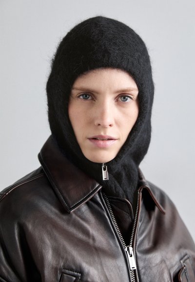 Balaclava neagră din lână cu textură moale, purtată cu o jachetă din piele maro, dotată cu fermoar și guler. Fundal neutru.