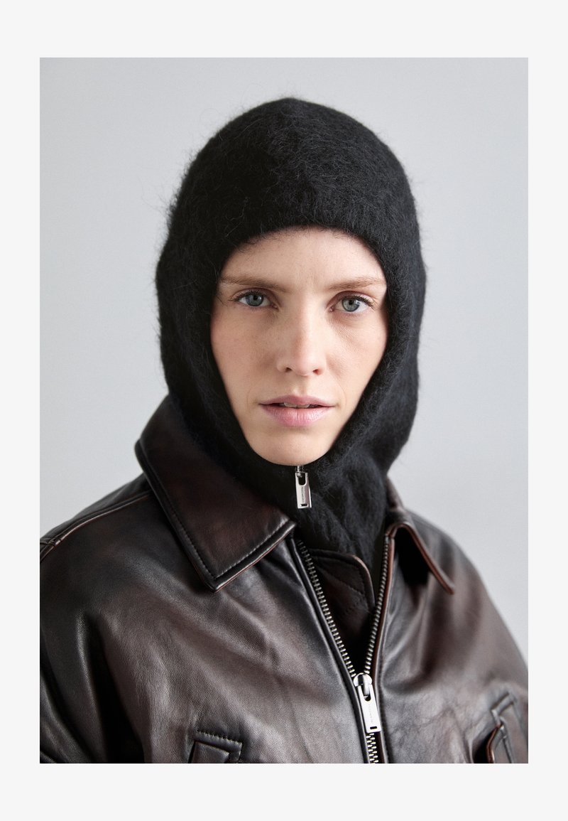 Fekete gyapjú balaclava, puha textúrával, barna bőrkabáttal párosítva, melyen cipzár és gallér található. Semleges háttér.