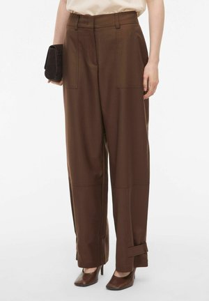 Bruine wijde broek gemaakt van een gladde stof, voorzien van voorzakken en opgerolde boorden. Gecombineerd met bruine hakken.